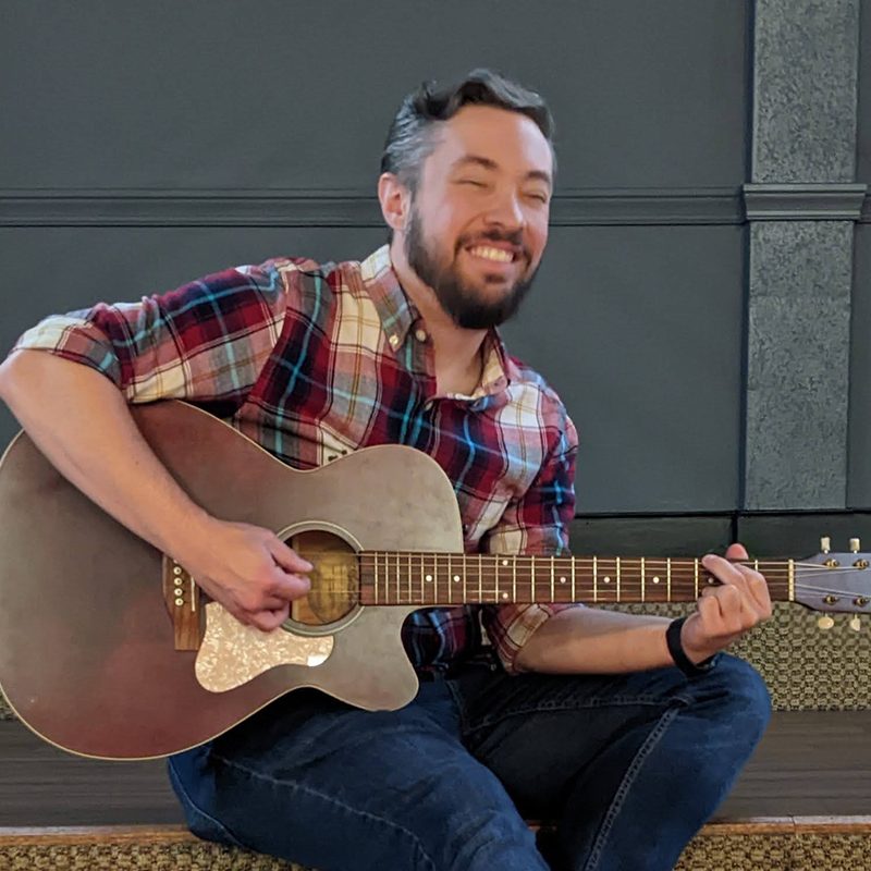 Chris LaFay sitting cross-legged with an acoustic guitar, laughing